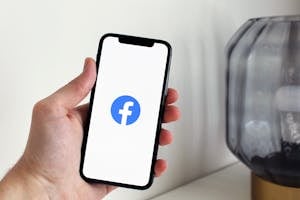 Close-up of a hand holding a smartphone displaying the Facebook logo indoors.