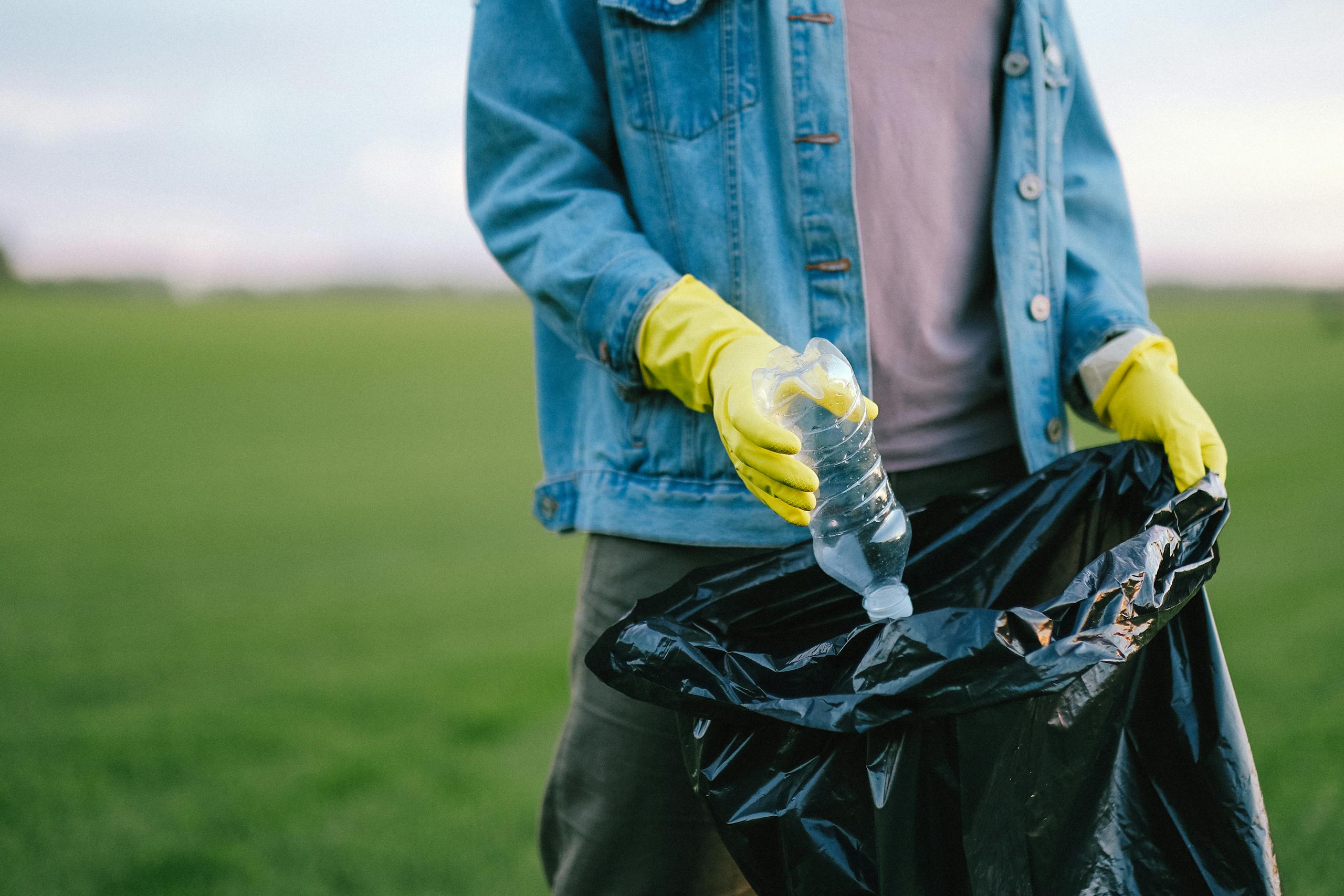 Volunteer in denim jacket and gloves picks up plastic bottle into trash bag in green field.