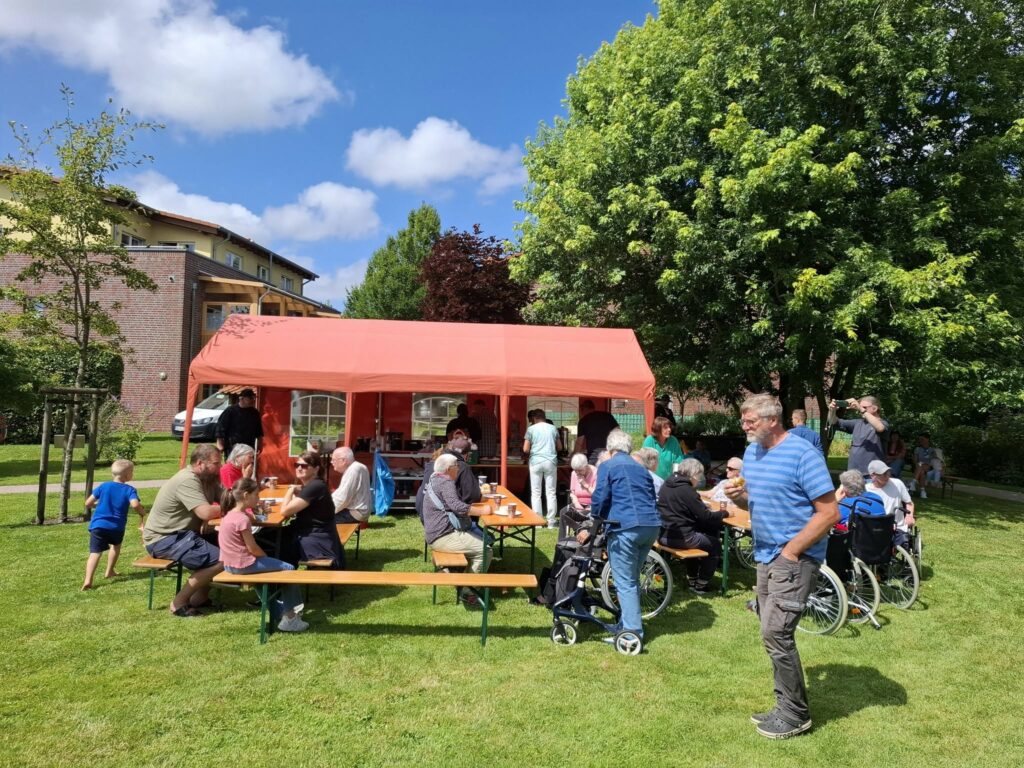 Besucherinnen und Besucher sitzen und essen gemeinsam im Verpflegungsbereich des Spielefests 2026 in Hooksiel, während Familien das sonnige Festgelände im Park genießen.