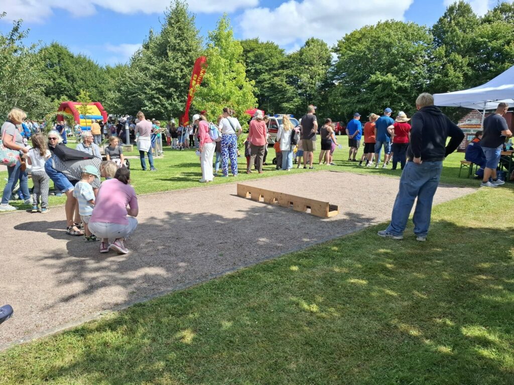 Familien und Besucher verfolgen ein Wurfspiel auf einer Mitmachfläche beim Spielefest 2026 in Hooksiel, während zahlreiche Aktionsstände und eine Hüpfburg das sonnige Festgelände im Park beleben.