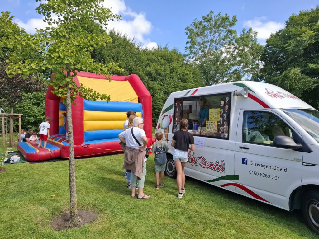 Kinder spielen auf einer Hüpfburg und Besucher stehen am Eiswagen beim Spielefest 2026 in Hooksiel, während Familien das sonnige Festgelände im Park genießen.