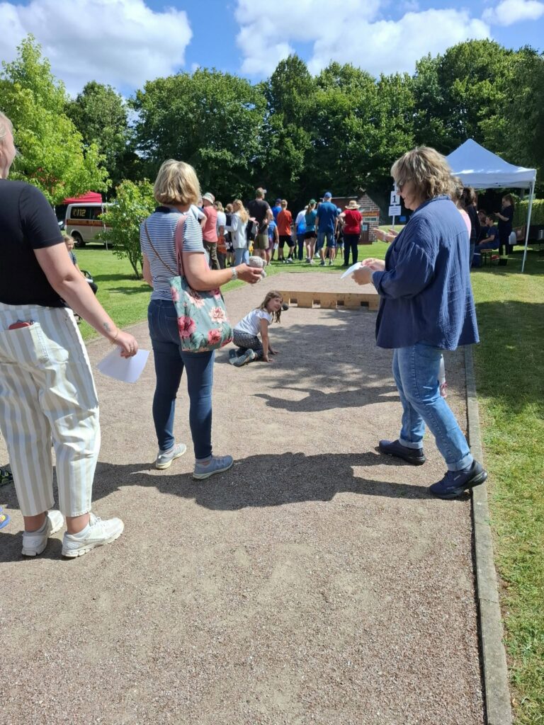 Kinder und Erwachsene nehmen an einer Mitmachstation mit Wurfspiel beim Spielefest 2026 in Hooksiel teil, während sich weitere Besucher auf dem sonnigen Festgelände aufhalten.