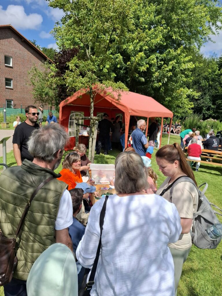 Kinder und Erwachsene nehmen an einer Bastel- und Mitmachstation beim Spielefest 2026 in Hooksiel teil, während zahlreiche Besucher das sonnige Festgelände im Park genießen.